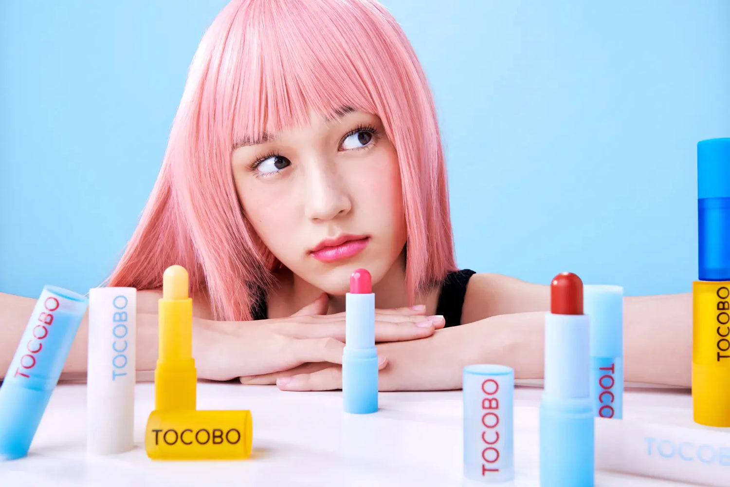 Young woman with pink hair surrounded by colorful lip balms and lipsticks in Tocobo collection
